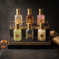 Six small cosmetic bottles with ornate gold caps and decorative labels are arranged on a black-and-gold display stand. The bottles contain pale yellow, amber, pink, green, blue-gray, and off-white liquids, with one bottle showing a gold glittery product.