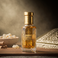 A clear glass perfume bottle filled with amber liquid, featuring a gold cap and a gold geometric label that reads 'Finashik Misk.' The bottle is set on a wooden surface with a warm, atmospheric background including a bowl of white flowers, a woven textile, and soft smoke.
