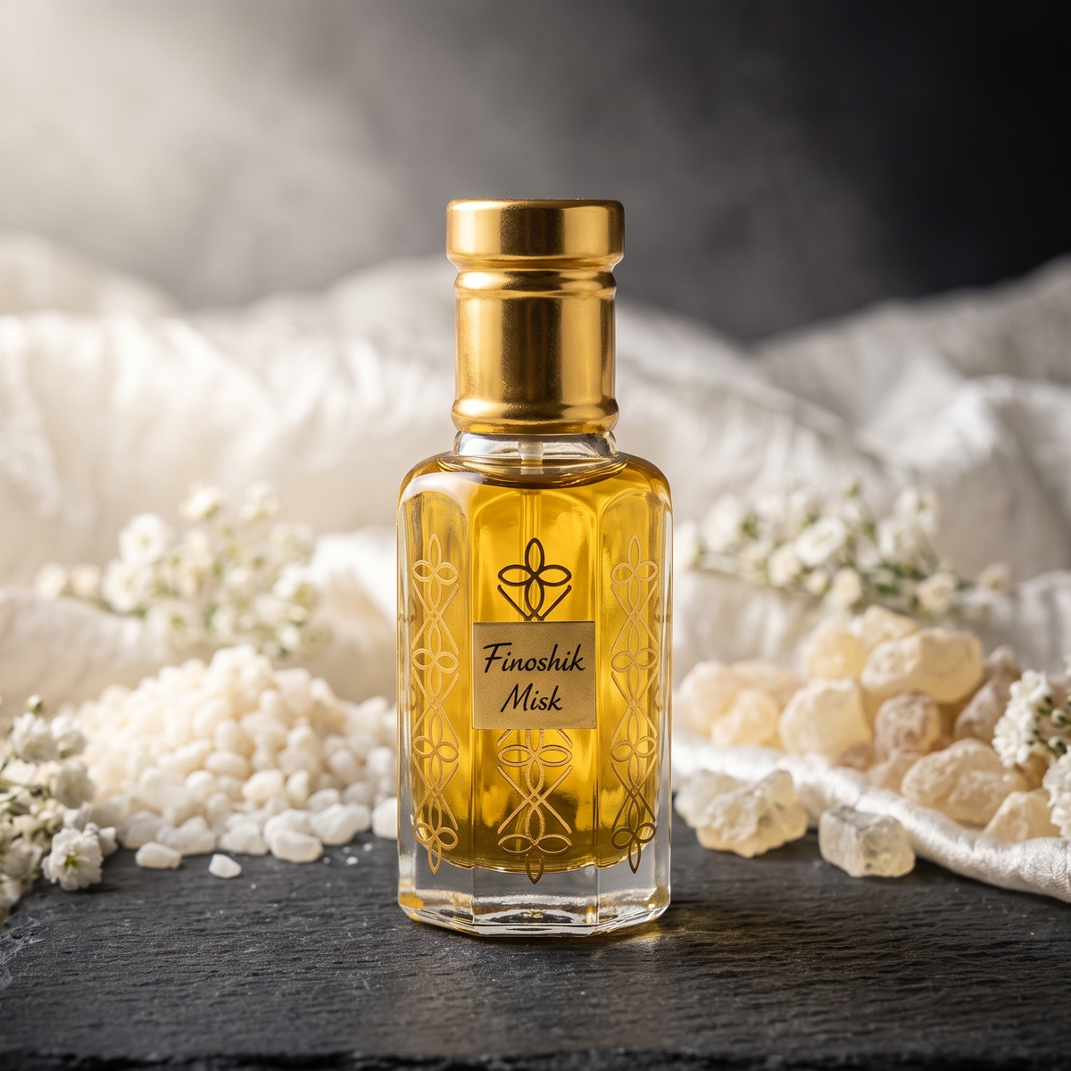 A small clear glass bottle filled with golden yellow liquid, topped with a metallic gold cap, standing on a dark slate board. The bottle has ornate gold etched patterns and a label reading 'Finashik Mask.