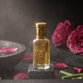 A small rectangular glass perfume bottle with a gold cap stands on a dark pedestal. The bottle contains a golden liquid and has ornate gold detailing on the label.